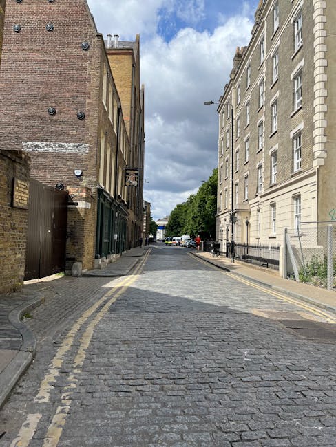 A narrow residential street in Lambeth with cobblestone paving, flanked by multi-storey buildings on both sides. On the left, there are brick buildings with some windows and a dark wooden gate, while on the right, a large cream-colored building features multiple windows and decorative architectural elements. Several vehicles are parked at the end of the street, and the sky above is partly cloudy with patches of blue. This scene illustrates a typical setting where home relocation and furniture transport might require careful navigation through restricted or narrow streets, with visible traffic management and potential access challenges for moving services such as those provided by Movers Lambeth.