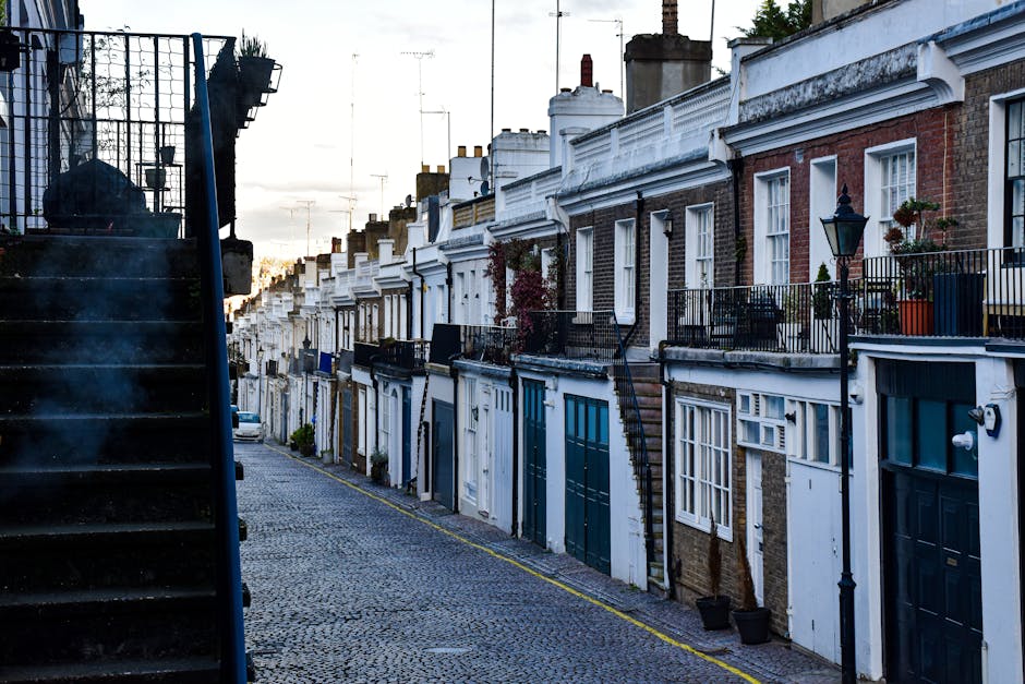 A street scene in Lambeth featuring a row of terraced houses with brick and white-painted facades, each with small front gardens or balconies enclosed by black iron railings. Several plants and potted flowers are visible on the balconies. The pavement is cobbled, and a narrow road runs along the front of the houses. On the left side of the image, a black outdoor staircase with metal steps and a railing leads to an upper floor of one of the buildings. The street appears quiet with no people in view, and the lighting suggests early morning or late afternoon. This setting is typical for house transfers and relocation services, with the narrow street and residential architecture reflecting the challenges of furniture transport and home removals in confined urban areas, which [COMPANY_NAME] handles with care.