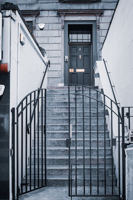 A set of concrete stairs leading up to a dark grey front door with a large windowpane divided into smaller sections at the top of the steps. The stairs are flanked by white walls with black metal railings on both sides, creating a secure boundary around the staircase. The door has a modern design with a letterbox and doorbell, and the surrounding building features grey stonework. There is no visible signage or vehicles in the image. The scene appears well-lit, capturing the exterior entrance of a residential property suitable for house removals and furniture transport, with the staircase potentially used during moving logistics involving stairs and lifts, as advised for SW2, Lambeth.