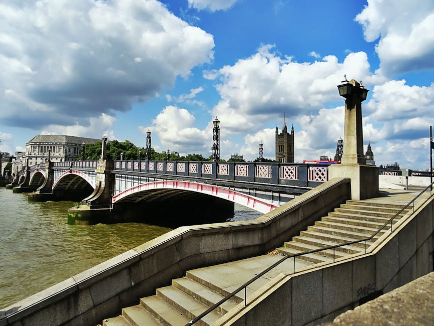 A stone staircase with metal handrails descending from a riverside promenade towards the River Thames, with a decorative railing and lamp posts lining the walkway. The bridge arching over the river features a classic design with white and red accents, supported by stone piers. In the background, there are historic buildings, including the Parliament Tower with its distinctive Gothic architecture, under a partly cloudy sky. At the riverside, cardboard boxes, plastic wrap, and blankets are visible, indicating a home relocation process managed by moverslambeth.co.uk, who are preparing for furniture transport and loading operations near the bridge.