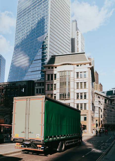 A large moving truck with a green cargo cover is parked on a city street, next to a modern high-rise building with reflective glass windows and a historic-style building with beige stone façade and multiple rectangular windows. The truck is positioned close to the curb, with its rear doors closed, and there are a few pedestrians and cyclists visible nearby. The scene is well-lit with clear weather, and the buildings around suggest an urban environment suitable for home relocation or furniture transport services, as provided by Movers Lambeth. The truck's placement on the street indicates loading or unloading activity, integral to the moving process involving packing, transportation, and delivery within the city.