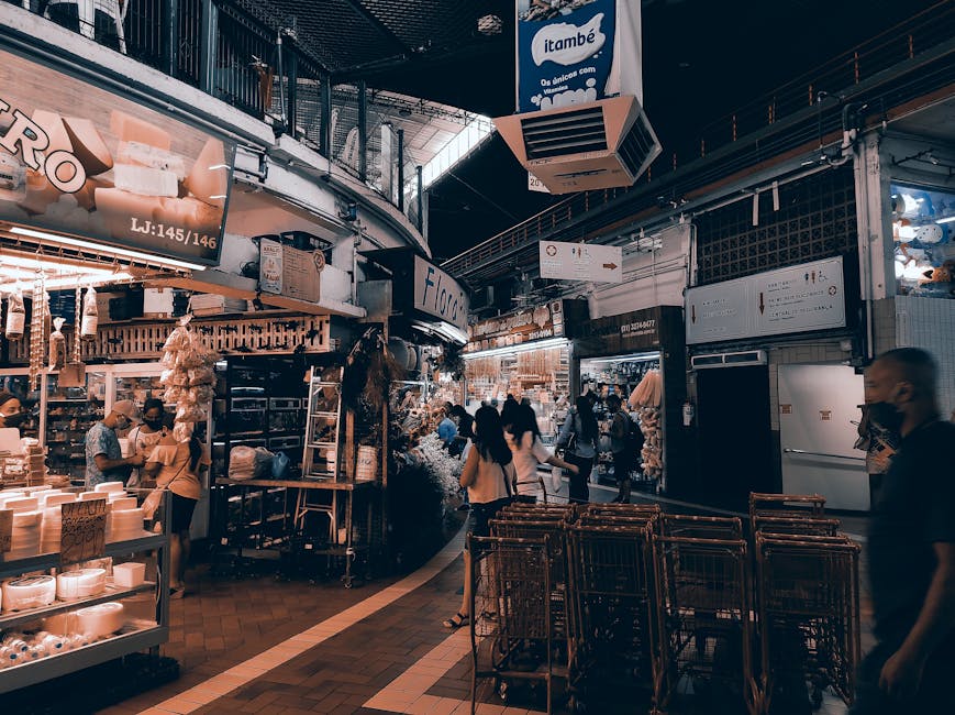 Interior view inside a busy market hall with various retail stalls selling goods, including packaged food, household items, and textiles, with some products displayed on shelves and racks. Several shoppers are walking through the aisle, some carrying shopping baskets. The ceiling has large air conditioning units and hanging signs indicating different sections. The market's lighting is bright, illuminating the merchandise and creating a vibrant atmosphere. The floor is tiled, and the scene suggests a typical setting for house removals or packing activities when relocating household items from such a market environment. The image also shows a partially visible rear section of a van or storage unit, hinting at the loading or unloading process carried out by [COMPANY_NAME], specializing in removals and home relocation services, with a focus on moving goods safely and efficiently in narrow or confined spaces.