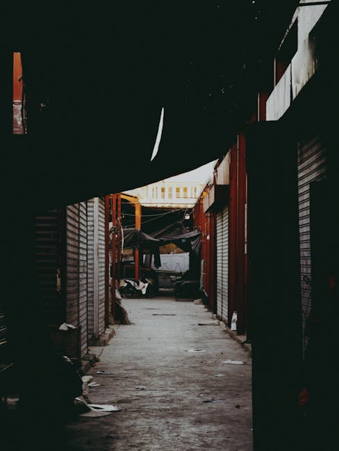 A narrow, enclosed alleyway between tightly packed market stalls or storage units with wooden and metal paneling, partially covered by hanging fabric and tarpaulins. The ground is concrete with scattered debris and small items, and at the far end, there is a glimpse of a brighter space with possible outdoor elements. While no furniture or moving equipment is visible in this image, this environment suggests a challenging access area where a professional removals service such as Movers Lambeth may need to assist with home relocation or furniture transport through confined urban spaces, especially relevant for jobs involving narrow access in Lambeth, as indicated by the page title. The image captures the logistical complexity of moving within tight city locations, emphasizing the need for careful planning during packing and moving operations.