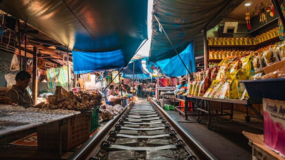 Interior view inside a busy market hall with various retail stalls selling goods, including packaged food, household items, and textiles, with some products displayed on shelves and racks. Several shoppers are walking through the aisle, some carrying shopping baskets. The ceiling has large air conditioning units and hanging signs indicating different sections. The market's lighting is bright, illuminating the merchandise and creating a vibrant atmosphere. The floor is tiled, and the scene suggests a typical setting for house removals or packing activities when relocating household items from such a market environment. The image also shows a partially visible rear section of a van or storage unit, hinting at the loading or unloading process carried out by [COMPANY_NAME], specializing in removals and home relocation services, with a focus on moving goods safely and efficiently in narrow or confined spaces.
