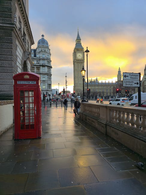 A street scene in central London during sunset, featuring a classic red telephone booth positioned on the sidewalk near a historic building facade. In the background, the iconic Big Ben clock tower and the Houses of Parliament are visible, illuminated by the setting sun. Pedestrians are walking along the wet pavement, some carrying packages or bags, with streetlights casting a warm glow. A row of parked cars and vehicles driving on the road can be seen, alongside a directional sign indicating routes to Trafalgar Square and Lambeth Bridge. The scene underscores the busy atmosphere of the capital, with potential relevance to home relocation or moving services offered by moverslambeth.co.uk, as part of the city’s bustling environment.