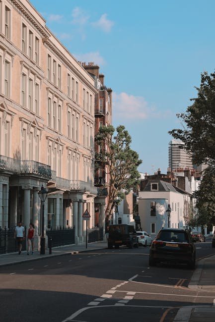 A street scene in Clapham Common, Lambeth, showing a row of multi-storey residential buildings with classic architectural features such as tall windows, balconies with wrought iron railings, and decorative facades. The street is paved and lined with mature trees providing shade, with parked cars along the roadside including a black van positioned near the curb. In the background, there is a modern high-rise building contrasting with the older structures. The weather is clear with a blue sky and some scattered clouds, indicating daytime. The presence of pedestrians walking along the sidewalk suggests typical urban activity. This setting exemplifies the typical environment for house removals and furniture transport services, involving loading maneuvered by professional movers like [COMPANY_NAME], preparing residential properties for home relocation or packing and moving processes, with vehicles possibly visible nearby for efficient relocation logistics.