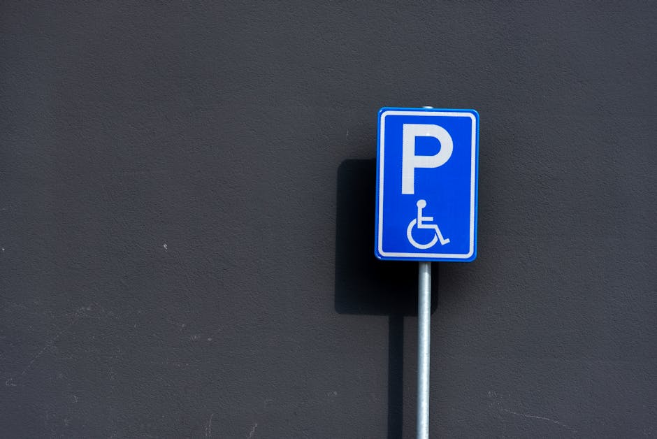 A blue parking sign featuring a white letter 'P' and a wheelchair symbol indicating accessible parking, mounted on a silver metal pole against a dark grey wall. The shadow of the sign is cast onto the wall, suggesting outdoor lighting. This signage is positioned near a property entrance or pavement area, commonly used for designated parking for disabled individuals or special access purposes. The image emphasizes the importance of accessible parking permits for house removals and furniture transport services, highlighting the need for compliance with local parking regulations during home relocation or moving processes. Occasionally, Movers Lambeth may assist clients with parking permit requirements or navigating local parking restrictions for removals vans, especially in areas like Lambeth where parking regulations are strictly enforced, as explained on their website about council parking permits.