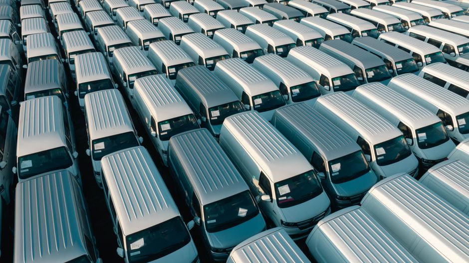 An aerial view of a large parking lot filled with white commercial vans arranged in neat rows, each equipped with metal roof racks and visible windshields. The vans are parked close together on a paved surface, with sunlight casting shadows on the vehicles. This scene illustrates a fleet of vehicles likely used for home relocation and furniture transport services by Movers Lambeth, positioned in preparation for a logistics operation. The image emphasizes the scale of vehicle coordination involved in household removals, highlighting the importance of parking permits and logistical planning for moving companies operating in Lambeth, as discussed on the Movers Lambeth website.