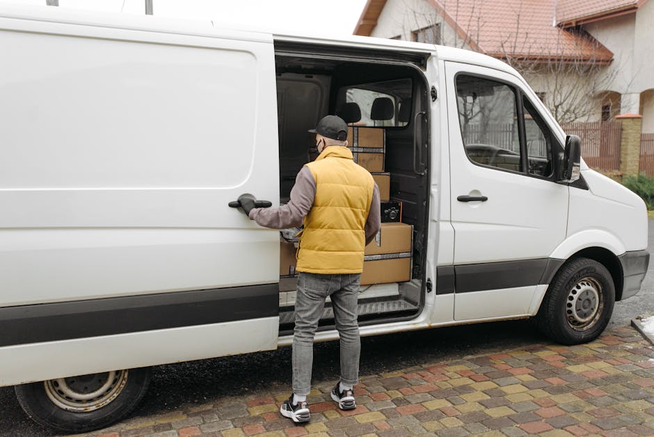 A person wearing a yellow padded vest, grey hoodie, and grey jeans is standing outside a white commercial van, loading or unloading cardboard boxes from the side door. The van is parked on a brick-paved driveway, with the sliding door open to reveal stacked boxes of various sizes secured with black straps inside. In the background, there are residential buildings with pitched roofs, trees, and a fence, indicating a suburban residential area. The scene is illuminated by natural daylight, capturing the process of home relocation or furniture transport as part of a professional removal service, consistent with house removals in Lambeth. Movers Lambeth specialize in packing and moving logistics, including loading and unloading in tight or accessible spaces.