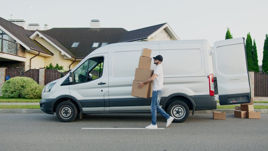A young man wearing a white t-shirt, blue jeans, white sneakers, and a black cap is walking beside a white cargo van parked on a residential street, carrying three cardboard boxes stacked vertically in his arms. The van's sliding side door is closed, and three additional cardboard boxes are on the pavement nearby. The van is positioned in front of a modern detached house with a pitched roof, dormer windows, and a driveway, with a well-maintained garden featuring shrubs and trees. The scene depicts the process of home relocation, with the man actively involved in packing or furniture transport, supported by the van for moving logistics. The environment is lit naturally, indicating daytime, with a clear sky and residential area surroundings as part of a professional removals service by Movers Lambeth.