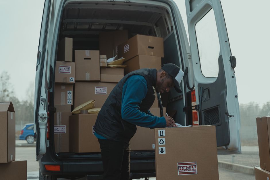 A person wearing a yellow padded vest, grey hoodie, and grey jeans is standing outside a white commercial van, loading or unloading cardboard boxes from the side door. The van is parked on a brick-paved driveway, with the sliding door open to reveal stacked boxes of various sizes secured with black straps inside. In the background, there are residential buildings with pitched roofs, trees, and a fence, indicating a suburban residential area. The scene is illuminated by natural daylight, capturing the process of home relocation or furniture transport as part of a professional removal service, consistent with house removals in Lambeth. Movers Lambeth specialize in packing and moving logistics, including loading and unloading in tight or accessible spaces.