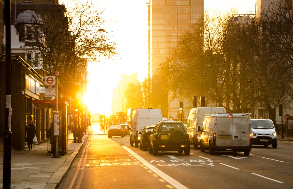 A busy urban street scene during sunset with vehicles including several white vans, small cars, and a bus parked along the roadside. The sidewalk features a bus stop shelter with people waiting, some walking, and various street fixtures such as lamp posts and bus signs. Tall modern buildings and leafless trees line the street, illuminated by warm, golden sunlight casting long shadows. The image captures a typical day in Lambeth, illustrating the urban environment where house removals and furniture transport services by Movers Lambeth are often required for home relocation or moving logistics, with the loading process likely happening nearby for ongoing home removals projects.