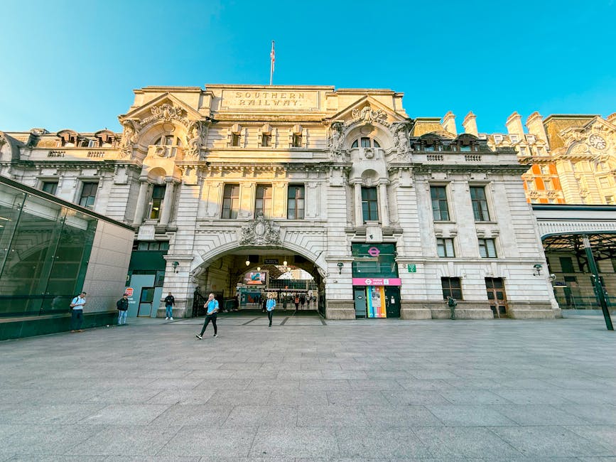 A wide view of Vauxhall Station, showcasing its historic stone facade with intricate architectural details and a large central archway leading into the station entrance. The building features ornate carvings and decorative elements on the upper levels, with a sign indicating 'Southern Railway' at the top. In front of the station, there is a spacious paved area where multiple individuals are walking, some carrying backpacks or bags, and others waiting near the entrance. To the left, a modern glass addition reflects the historic architecture. Inside the archway, a few people are visible near the station entrance, while outdoor equipment such as trolleys and luggage carts are positioned nearby. This scene captures the typical environment for local home relocation or furniture transport services, emphasizing the station as a key point in the moving logistics process, with natural daylight illuminating the scene and clear skies overhead.
